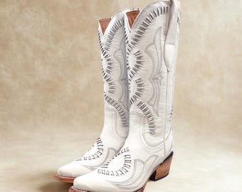 Könnte beinhalten: Ein Paar weiße Cowboy-Stiefel mit aufwendigen Nähten und Aussparungen. Die Stiefel haben eine spitze Zehenpartie, einen gestapelten Absatz und eine hellbraune Sohle. Der hohe Schaft hat dekorative Nähte und eine Zugschlaufe.