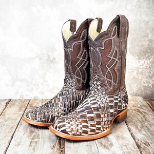 Puede incluir: Un par de botas de vaquero de cuero marr&oacute;n. El pie presenta un dise&ntilde;o tejido, mientras que la ca&ntilde;a superior tiene un patr&oacute;n cosido decorativo. Las botas tienen una puntera cuadrada y un tac&oacute;n apilado.