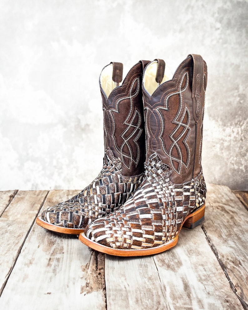 Puede incluir: Un par de botas de vaquero de cuero marr&oacute;n. El pie presenta un dise&ntilde;o tejido, mientras que la ca&ntilde;a superior tiene un patr&oacute;n cosido decorativo. Las botas tienen una puntera cuadrada y un tac&oacute;n apilado.