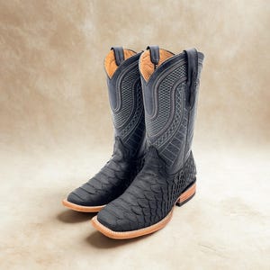 May include: A pair of black cowboy boots with a textured, scaly pattern on the foot and a smooth, stitched design on the upper shaft. The boots have a square toe and a light brown sole. The interior is a light tan color.