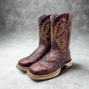May include: A pair of brown leather cowboy boots with intricate floral and scrollwork designs. The boots have a square toe, a light-colored sole, and decorative stitching in gold and cream colors.