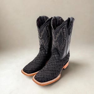 May include: A pair of black cowboy boots with a textured pattern on the foot and a smooth leather upper. The boots have a square toe and are accented with white stitching. The soles are a light brown color.