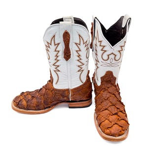 May include: A pair of cowboy boots with a unique design. The boots feature a white upper with brown decorative stitching and a textured, brown lower section. The boots have a square toe and a stacked heel.
