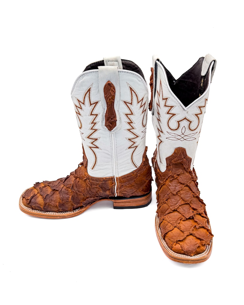 May include: A pair of cowboy boots with a unique design. The boots feature a white upper with brown decorative stitching and a textured, brown lower section. The boots have a square toe and a stacked heel.