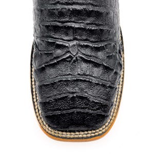 May include: Close-up of a black leather cowboy boot. The boot features a square toe and a textured, crocodile-like pattern. The sole is tan with a stitched border.