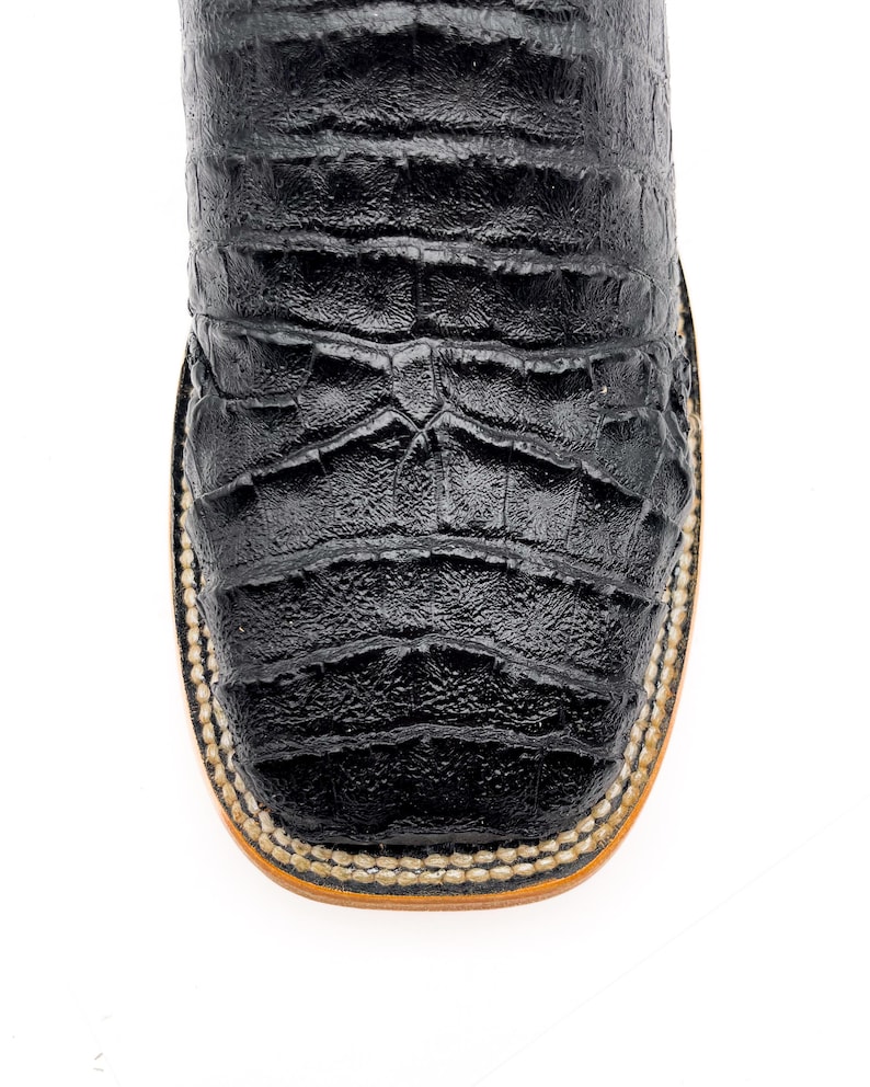 May include: Close-up of a black leather cowboy boot. The boot features a square toe and a textured, crocodile-like pattern. The sole is tan with a stitched border.