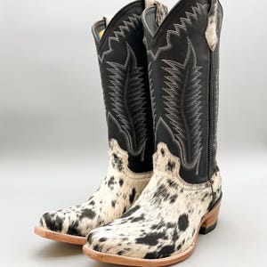 May include: A pair of black and white cowhide cowboy boots with a black leather shaft and intricate stitching. The boots have a pointed toe and a low heel.