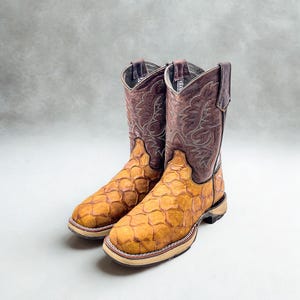 May include: A pair of cowboy boots with a unique design. The boots feature a brown leather upper with intricate stitching and a textured, golden-brown foot. The soles are light-colored with a brown and red trim.