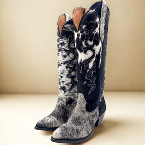 May include: A pair of tall cowboy boots with a black and white spotted pattern and black accents. The boots have a pointed toe and a stacked heel. The interior is a light brown color.