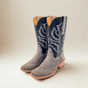 May include: A pair of cowboy boots with a gray suede foot and a black leather upper. The boots feature intricate white stitching and a square toe design. The heels are a light brown color.