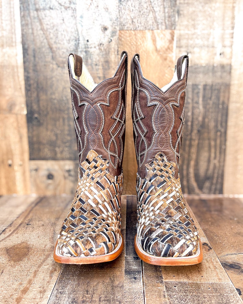 Puede incluir: Un par de botas de vaquero de cuero marr&oacute;n. El pie presenta un dise&ntilde;o tejido, mientras que la ca&ntilde;a superior tiene un patr&oacute;n cosido decorativo. Las botas tienen una puntera cuadrada y una suela marr&oacute;n claro.