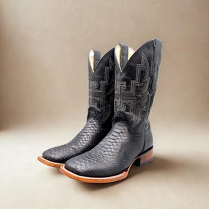 May include: A pair of black cowboy boots with a snakeskin pattern on the foot and a decorative stitched design on the shaft. The boots have a square toe and a brown sole.