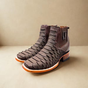 May include: A pair of brown leather boots with a unique scale-like texture on the front. The boots have a square toe, a white sole with an orange trim, and a brown heel. The ankle area is a darker brown leather.