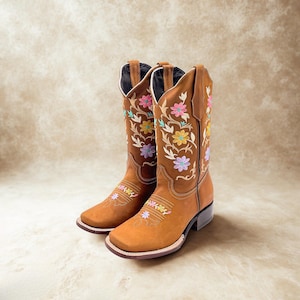 May include: A pair of brown cowboy boots with square toes. The boots are adorned with colorful floral embroidery in pink, yellow, and purple. The boots have a stacked heel and a leather sole.