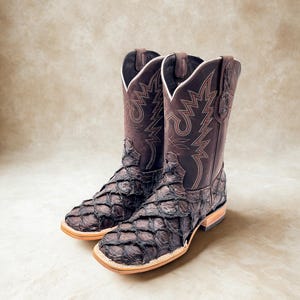May include: A pair of brown cowboy boots with a unique scale-like texture on the foot and lower leg. The upper shaft features decorative stitching and a classic western design. The boots have a square toe and a light-colored sole.