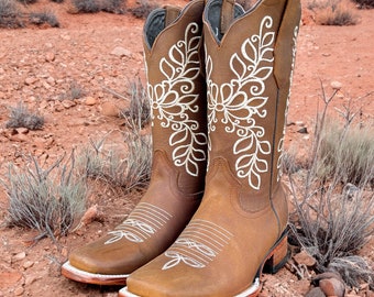 May include: A pair of brown leather cowboy boots with white floral embroidery. The boots have a square toe and a stacked heel. The boots are displayed outdoors on reddish-brown soil with sparse vegetation.