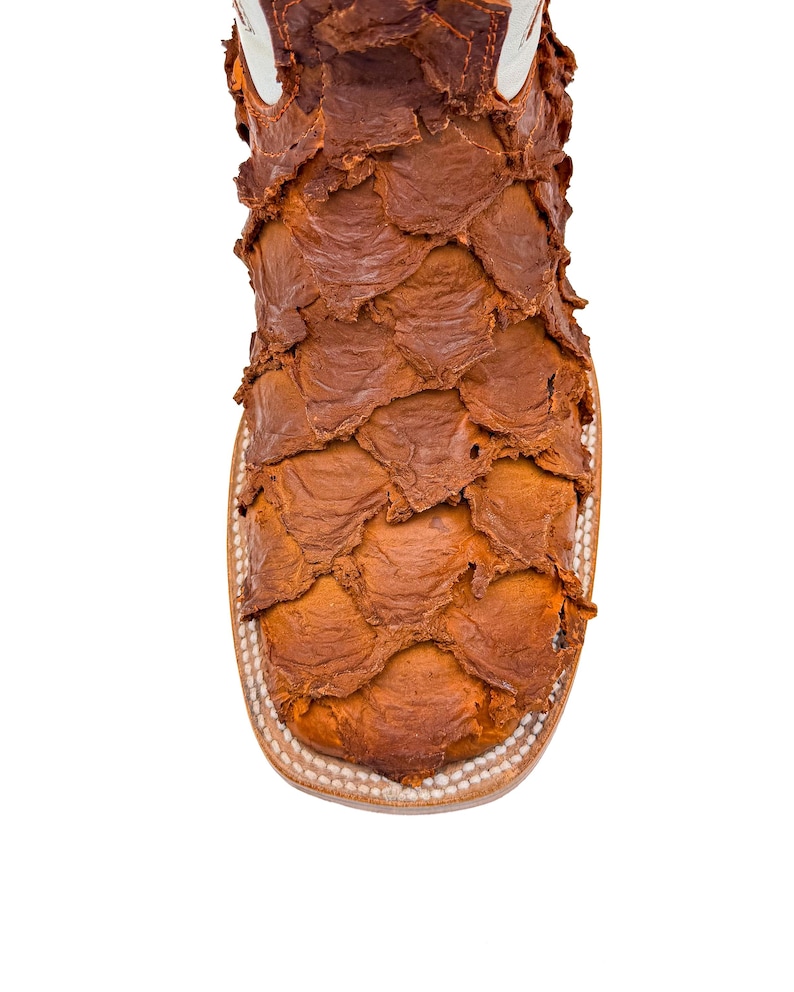 May include: Close-up of a brown leather boot with a unique, textured design. The boot features a square toe and a white upper. The leather has a scale-like pattern, creating a distinctive look. The boot's construction is visible.