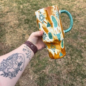 Könnte beinhalten: Ein großer, isolierter Becher mit einem braunen und weißen Muster, türkisfarbenen Glitzerakzenten und einem türkisfarbenen Glitzergriff. Der Becher hat geometrische und Bullenschädel-Designs.