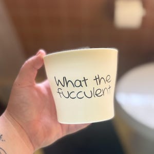 May include: A cream-colored ceramic cup with the words "What the fucculent" in black script. The cup is held in a hand, with a blurred background of a bathroom setting.