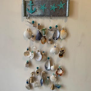 Peut inclure: Un carillon à coquillages avec une plaque en métal représentant une ancre et trois étoiles de mer. La plaque est peinte en bleu turquoise. Le carillon est composé de divers coquillages, perles et cloches.