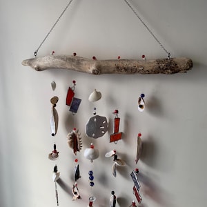 May include: A driftwood mobile featuring a variety of seashells, glass beads, and ceramic tiles. The mobile is suspended from a chain and has red, blue, and white accents.