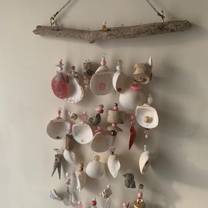 May include: A seashell wind chime with a driftwood hanger. The chime features various seashells, beads, and bells in shades of pink, white, and brown.