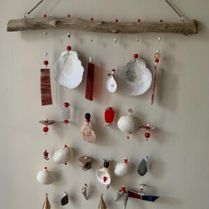 May include: A white driftwood wind chime with red beads and seashells hanging from it. The wind chime is made of natural materials and has a rustic look.