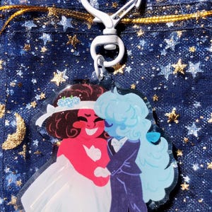 May include: A clear acrylic charm featuring two cartoon characters in wedding attire, one red and one blue. The red character wears a white dress and the blue character wears a suit. The charm has a white heart-shaped clasp.