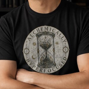 May include: A black short-sleeved t-shirt featuring a circular graphic on the chest. The design depicts an hourglass with celestial symbols and the text "ALCHEMICALLY HYPERFOCUSED" written in a circular border around the central illustration.