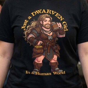 May include: A black short-sleeved t-shirt featuring a graphic illustration of a female dwarf with long brown hair and a beard, holding a wooden mug. The text "Just a DWARVEN Girl In a Human World" is printed in gold lettering around the character.