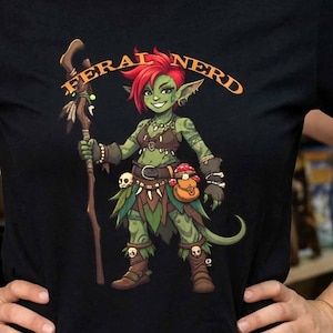 May include: A black short-sleeved t-shirt featuring a graphic illustration of a green-skinned goblin character with red hair holding a wooden staff. The text FERAL NERD is printed in an orange arc above the character, who wears brown leather gear.