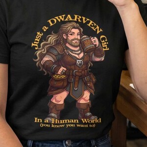 May include: A black t-shirt featuring a graphic illustration of a bearded female dwarf holding a wooden tankard. The text reads "Just a DWARVEN Girl In a Human World (you know you want to)" in gold lettering. The design is centred on the chest.