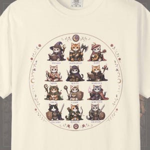 May include: A cream-colored t-shirt featuring a circular graphic of twelve cats dressed as fantasy role-playing characters. Each cat is labeled with its class: Wizard, Barbarian, Bard, Monk, Rogue, Warlock, Druid, Cleric, Sorcerer, Paladin, Ranger, and Fighter.