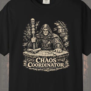 May include: A black t-shirt featuring a graphic print of a hooded figure in a cloak standing behind a stone table. The figure holds a staff topped with a skull. The text "CHAOS COORDINATOR" is printed in a bold, serif font below the table.