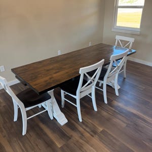 Puede incluir: Una mesa de comedor rectangular marrón oscuro con una base de pedestal blanca está rodeada por cuatro sillas blancas con asientos negros. Las sillas tienen un diseño de respaldo en forma de X. La mesa y las sillas están en una habitación con piso de madera.