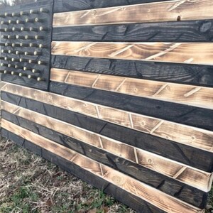May include: A wooden American flag with black and brown stripes. The flag is made of reclaimed wood and has a distressed look. The stars are made of metal and are attached to the flag with screws.