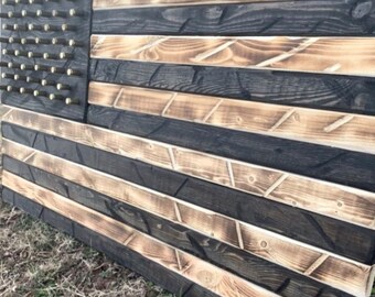 Op de afbeelding: Een Amerikaanse vlag van hout met zwarte en bruine strepen. De vlag is gemaakt van gerecycled hout en heeft een verweerde uitstraling. De sterren zijn van metaal en zijn met schroeven aan de vlag bevestigd.