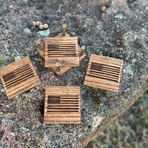May include: Set of four square wooden coasters with an engraved American flag design. The coasters are made of light brown wood and are arranged on a textured surface. The coasters are designed to hold a glass or mug.