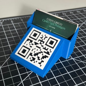 May include: A blue plastic business card holder with a white QR code on the front. The holder has a slot for a business card and is angled to display the card. The business card reads "Cathy Greco Cameo MacPherson Author C".