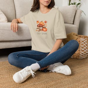 Puede incluir: Una camiseta beige de manga corta con un estampado gráfico de libros apilados, calabazas naranjas y hojas de otoño con el texto COZY SEASON impreso debajo. La camiseta se combina con pantalones vaqueros azules y zapatillas blancas.