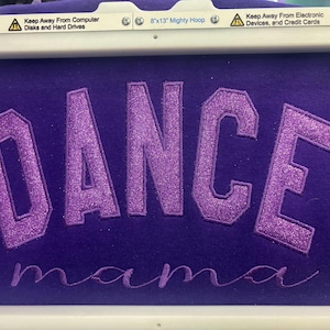 May include: Purple fabric with the word "DANCE" in large, glittery, purple letters, and "mama" in cursive below. The fabric is held in a white embroidery hoop. The top of the hoop has text that reads "Keep Away From Computer Disks and Hard Drives".