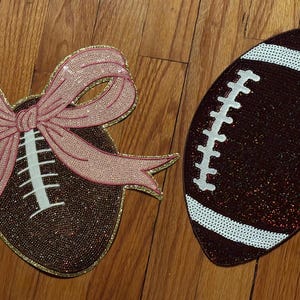 Könnte beinhalten: Zwei dekorative Football-förmige Artikel. Einer ist ein brauner, paillettenbesetzter Football mit weißen Nähten. Der andere ist ein brauner, paillettenbesetzter Football mit einer rosa Pailletten-Schleife. Beide haben einen goldenen Rand.