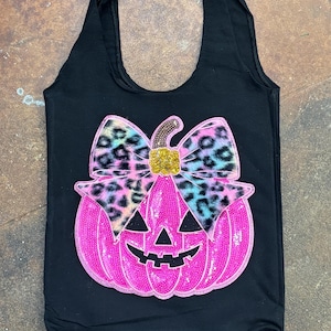 Puede incluir: Un bolso tote negro que presenta una gran calabaza de Halloween de color rosa brillante cubierta de lentejuelas. La calabaza está rematada con un lazo prominente con estampado de leopardo multicolor y un tallo central de purpurina dorada.