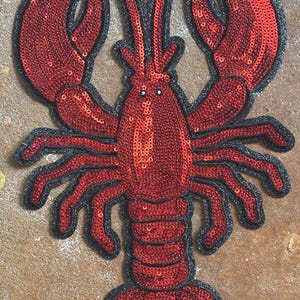 May include: A decorative lobster patch featuring vibrant red sequins covering the body and large claws. The crustacean design is outlined with a thick black border, highlighting the segmented tail, multiple legs, and two long antennae extending upwards.
