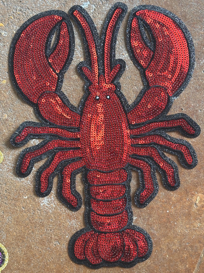 May include: A decorative lobster patch featuring vibrant red sequins covering the body and large claws. The crustacean design is outlined with a thick black border, highlighting the segmented tail, multiple legs, and two long antennae extending upwards.