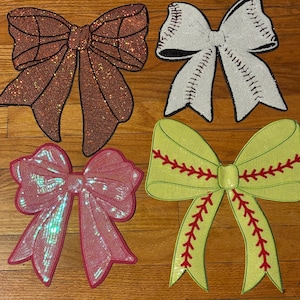 May include: Four decorative bows in the image. One is brown with a basketball design, another is white with baseball stitching, a pink sequined bow, and a green bow with red stitching. The bows are on a wooden surface.