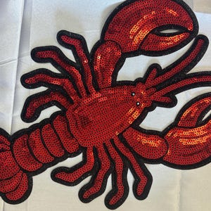 May include: A lobster-shaped patch covered in bright red sequins with a black stitched border. The design features two large claws, multiple legs, and a segmented tail, all rendered in a shimmering, reflective sequined material.