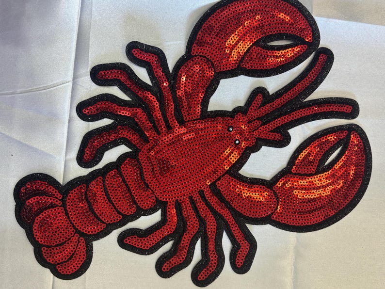 May include: A lobster-shaped patch covered in bright red sequins with a black stitched border. The design features two large claws, multiple legs, and a segmented tail, all rendered in a shimmering, reflective sequined material.