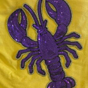 May include: A lobster-shaped patch featuring purple sequins and beads with a gold glitter border, attached to a solid yellow fabric background. The design includes two large claws, multiple legs, and antennae, all rendered in a vibrant purple colour.