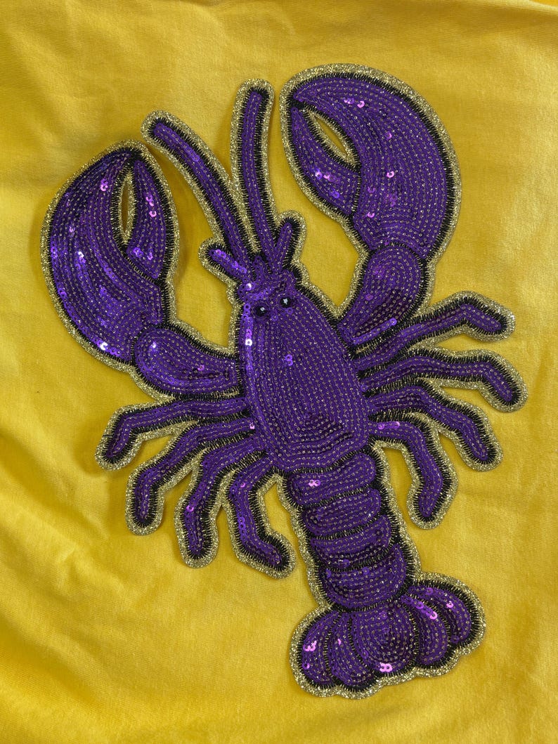 May include: A lobster-shaped patch featuring purple sequins and beads with a gold glitter border, attached to a solid yellow fabric background. The design includes two large claws, multiple legs, and antennae, all rendered in a vibrant purple colour.